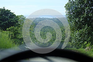 Road through jungle, view behind windshield of car. Palm trees and sea in background