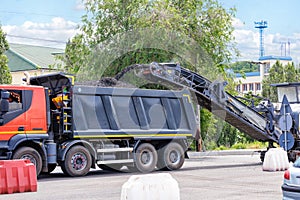 A road miller removes the top layer of old asphalt from a road section and loads it onto a dump truck