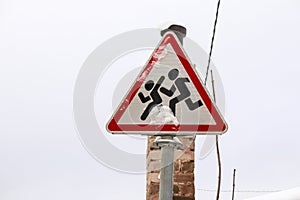 Road sign depicting running children, covered with snow against a white winter sky with a chimney behind it