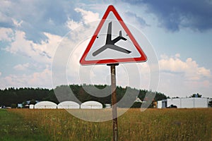 Road sign with red triangle and black plane going down landing standing in the field on blue sky background with clouds