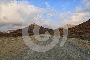 Road surrounded by hills covered in small rocks and sand under a blue cloudy sky