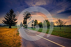 Road beside wheat field and cottage at sunset with colorful overcast sky..