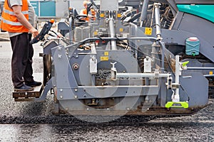 A road worker operates an asphalt paver and pours asphalt concrete while standing behind a control panel on a metal step on a
