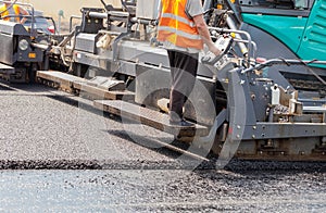 A road worker operates a tracked asphalt paver while paving fresh asphalt on the road on a summer day