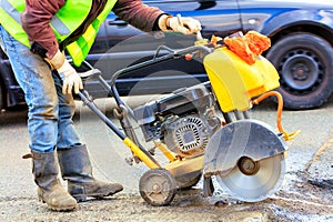 A road worker in reflective overalls uses a portable asphalt cutter to cut away worn asphalt with a diamond blade