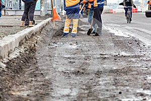 Road workers in orange and blue reflective overalls shovel debris into a pile on a road section being repaired