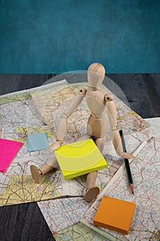 Roadmaps folded and scattered wooden manikin doll sitting holding a blank posted note