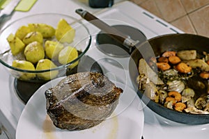 Roast beef lies on a plate next to a pan with stewed vegetables and a bowl of whole baked potatoes