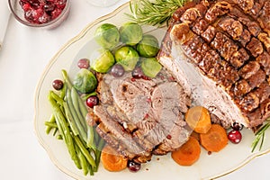 Roast pork with crust as Christmas ham with vegetables and cranberries on a white serving plate, holiday dinner, high angle view