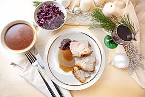 Roast pork slices with crust on a plate served on a festive table with sauce, red cabbage, wine glass, white candles and Christmas