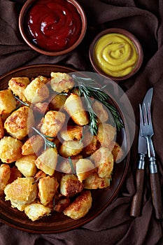 Roast Potatoes with spices and rosemary served on a clay dish with mustard and tomato sauce on an old wooden table, verticaL view