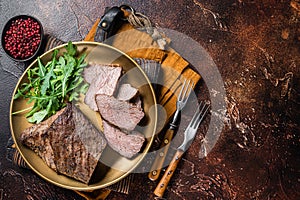 Roast Tri Tip or sirloin bottom beef steak on a plate with arugula. Dark background. Top view. Copy space