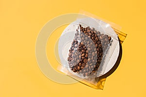 Roasted arabica coffee beans in blank transparent plastic bag with zipper on cement podium and yellow background