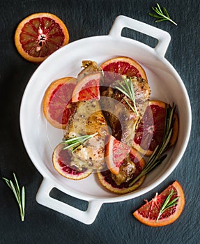 Roasted chicken legs on slices of red oranges in white baking dish. Black slate background.