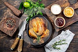 Roasted chicken with rosemary served on black plate with sauces on wooden table, top view.