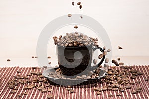 Roasted coffee beans falling down into black cup standing on black plate standing on tablemat.