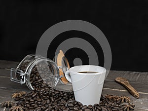 Roasted coffee beans in glass jar with a white coffee cup on vintage table.Closeup view with copy space on dark black background