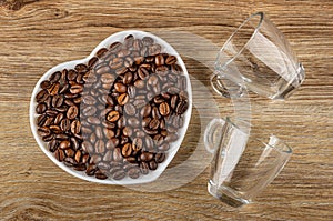 Roasted coffee beans in saucer in heart shape, empty transparent cups on wooden table. Top view