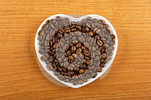 Roasted coffee beans in saucer in heart shape on wooden table. Top view