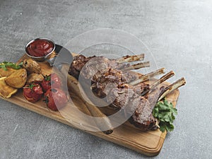 Roasted rack lamb chops on a wooden board with red sauce potatoes and tomatoes