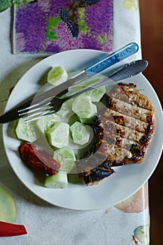 Roasted steak meat with bone, cucumber , ketchup fork and knife