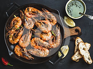 Roasted tiger prawns in iron grilling pan with fresh leek, lemon, bread and pesto sauce over black background