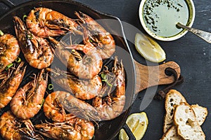 Roasted tiger prawns in iron grilling pan with fresh leek, lemon slices, bread and pesto sauce over black background