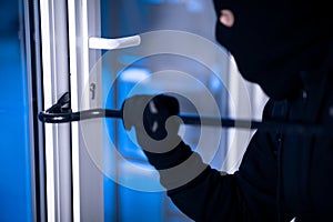 Robber in black balaclava cracking door with the crowbar