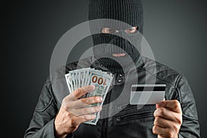 Robber, a thug in a balaclava holds a credit card in his hands on a dark background. Robbery, hacker, crime, theft. Copy space