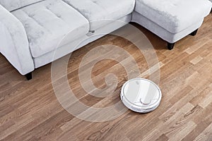 Robotic vacuum cleaner runs near sofa on laminate floor. Robot controlled by voice commands to direct cleaning. Modern smart
