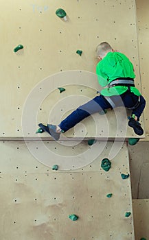 Rock climbing for children. Blond boy in a green shirt is climbing climbing wall. Bouldering class. Artificial track for safe