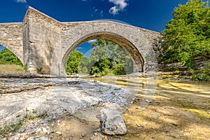 Rock stone bridge across small pond with green trees under blue sky. Historic scenery, idyll