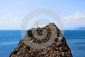 A rock texture of a mountainous rock on which a dried lone plant against the blue sea and sky. The peak of the mountain above sea