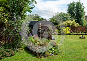 Rockery garden in the foreground, purple flowering buddleia tree to the left. Garden room with black and cedar cladding behind.