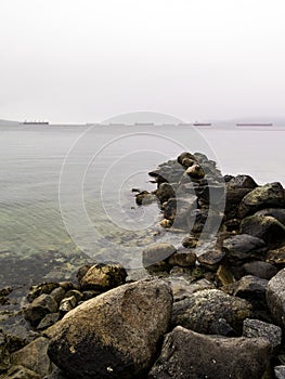 Rocks in the sea at stanley park seawall in vancouver with cargo ships at background in the fog