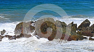 Rocks sticking out of the blue water at the shore, waves crashing on the rocks on a summer sunny day