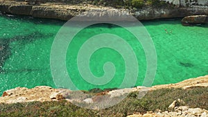 Rocky cliff at Cala Antena bay, calm green sea, two unrecognizable people swimming - typical Mallorca shore landscape