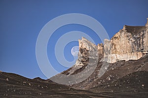 Rocky cliffs in the mountains rise with peaks against the background of the moon and the blue morning sky