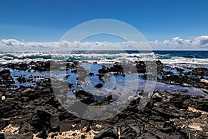 Rocky Hawaiian beach. Rocks, tidepool, sand. Wave and blue ocean in distance. Blue sky and clouds.