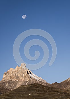 Rocky mountain peaks with the remains of snow against the background of a blue morning sky with a saturated moon