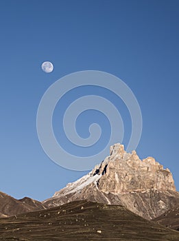 Rocky mountain peaks with the remains of snow against the background of a blue morning sky with a saturated moon