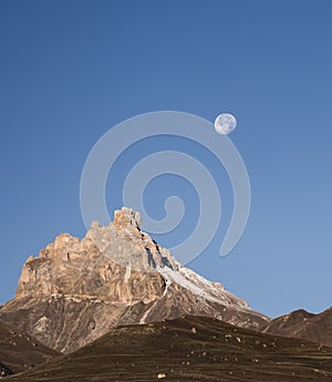 Rocky mountain peaks with the remains of snow against the background of a blue morning sky with a saturated moon