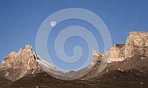 Rocky mountain peaks with the remains of snow against the background of a blue morning sky with a saturated moon