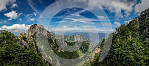 Rocky peaks and old pine trees cover the mountains under a bright blue sky with whispy clouds in Huangshan China