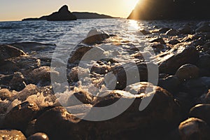 Rocky pebble sea beach with rocks at sunset, water with splashes hits the shore, close-up