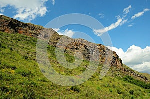 A rocky ridge of a high cliff running along the slope of a gentle hill under a summer cloudy sky