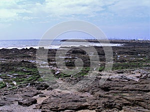 Rocky-rocky beach at low tide, in the blurred background of the Atlantic Ocean and a harbor entrance. Dark stones and water rests