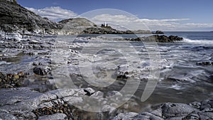 A rocky shoreline, with a white lighthouse on a headland. The beach is Bracelet Bay, Mumbles, south Wales