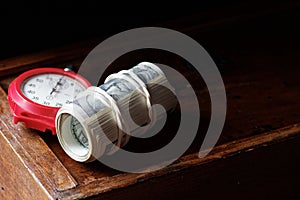 A roll of 100 american dollar banknotes with red clock stopwatch on wooden drawer on dark moody black background