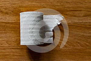 A roll of clean, plain toilet paper, cheap quality, dirty color, lies on a brown wooden table, slightly unwound